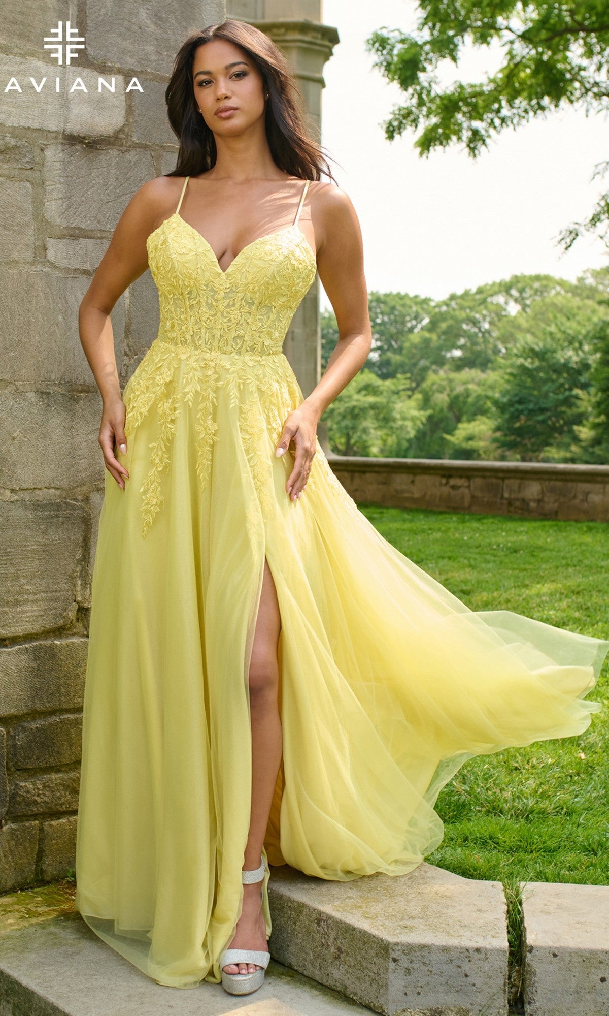 A woman wearing the Faviana 11438 long prom dress in yellow, featuring lace detailing and a thigh-high slit, stands outdoors by a stone wall.